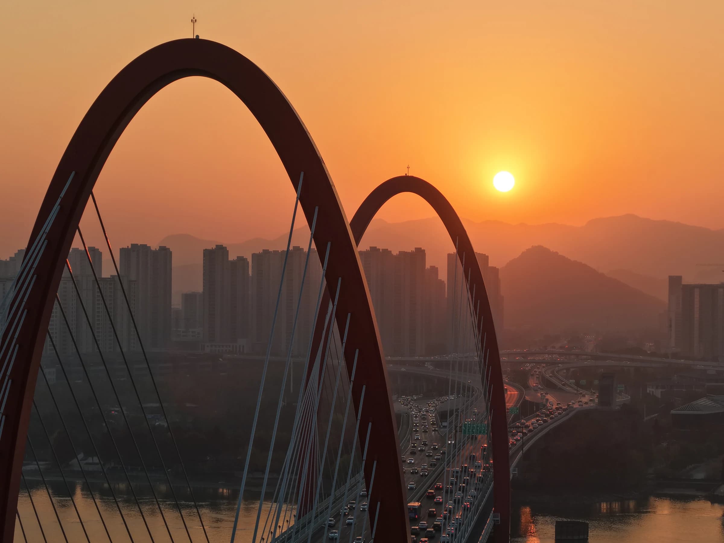 A sweeping arch bridge frames the setting sun above a distant city