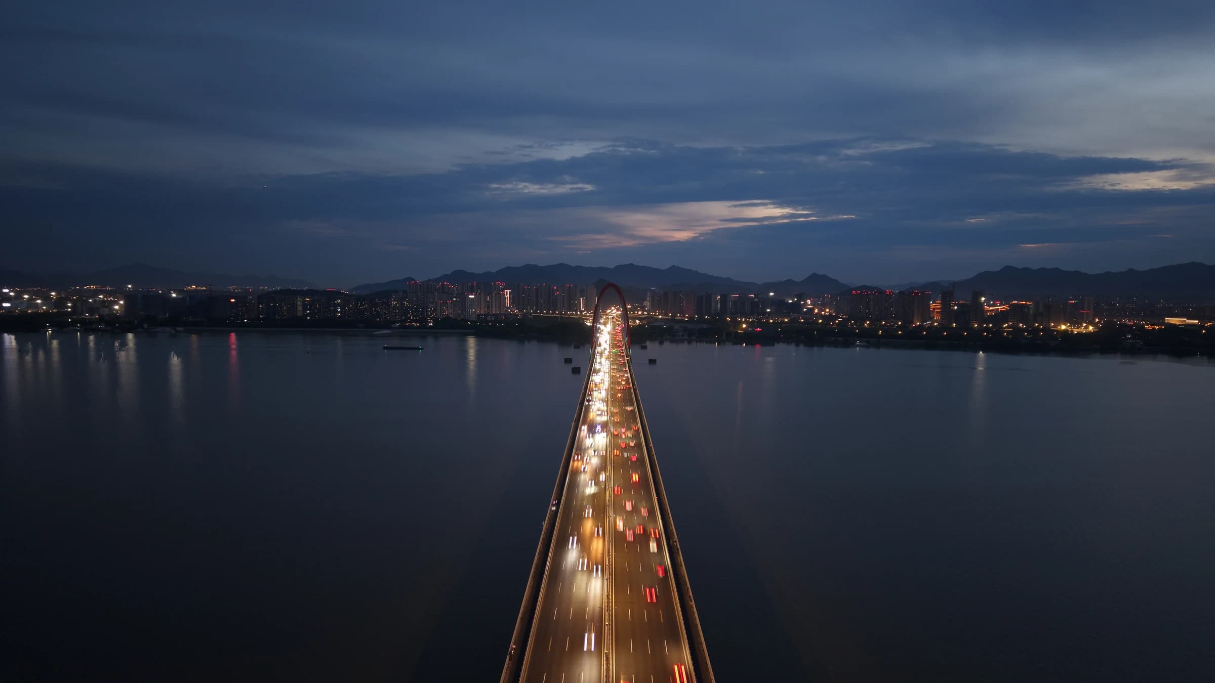 A stream of headlights crosses a long causeway over dark blue water at dusk