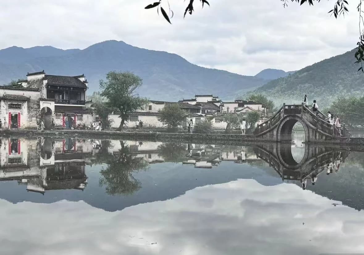 White-walled village houses and an arched stone bridge reflect in calm water beneath distant mountains